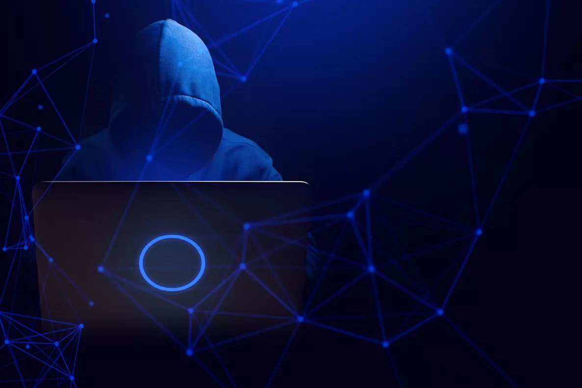 Fondo azul con negro un hombre con capucha frente a una computadora representando a un hackers y el Sodinokibi el ransomware más redituable para los hackers