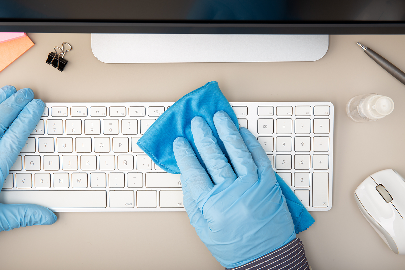 Teclado de computadora sobre escritorio con persona con guantes en sus manos limpiando el teclado porque se necesita de un plan de mantenimiento preventivo para TI