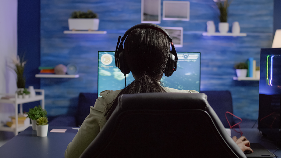 Mujer sentada de espaldas en una silla giratoria frente a una computadora con audífonos jugando en su cuarto good job el videojuego que te permite crear un caos en la oficina