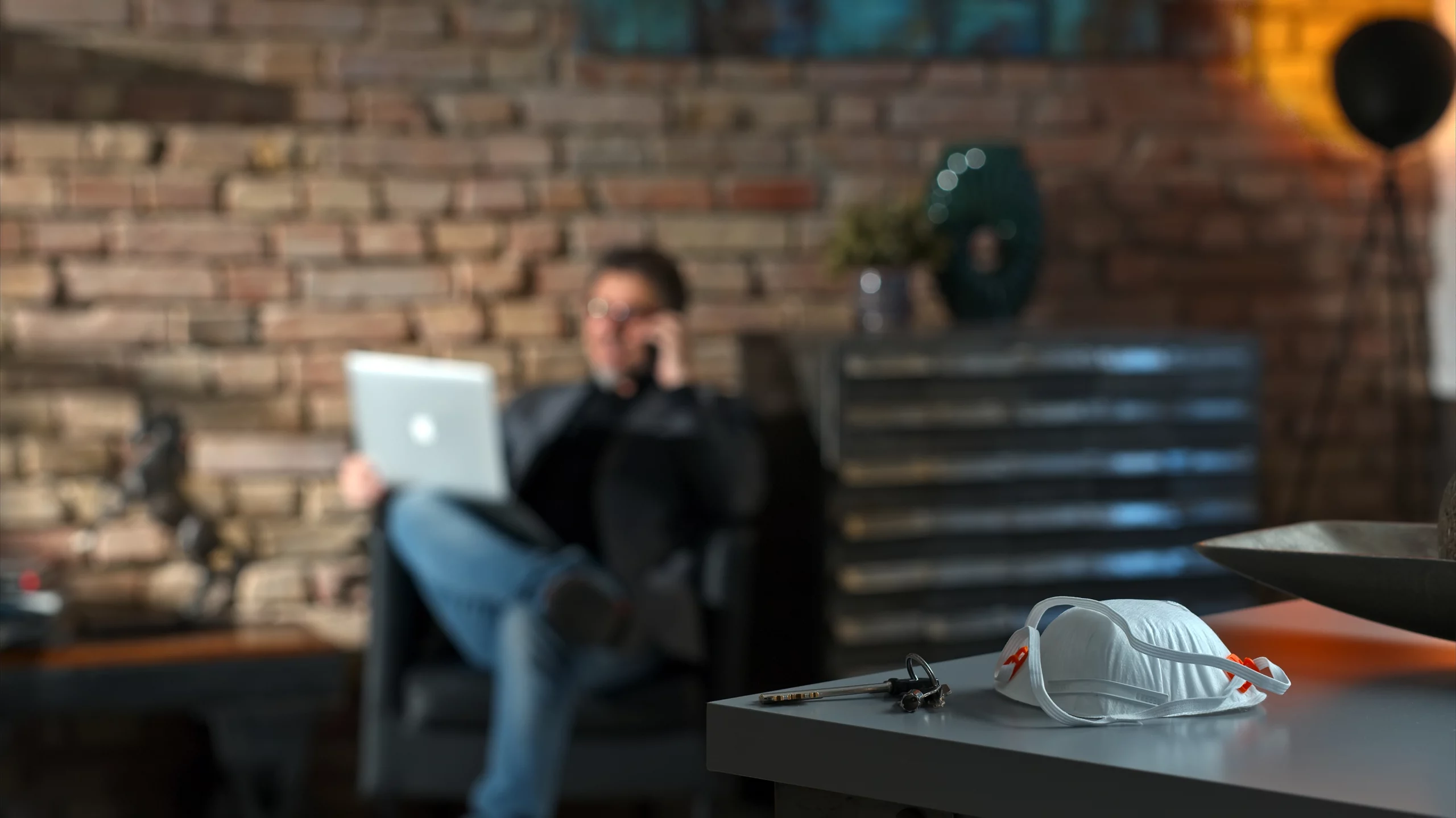 Hombre sentado en el sillón de su casa de fondo con una computadora, y en primer plano una mascarilla sobre una mesa representando cómo implementar el home office durante la contingencia por COVID-19