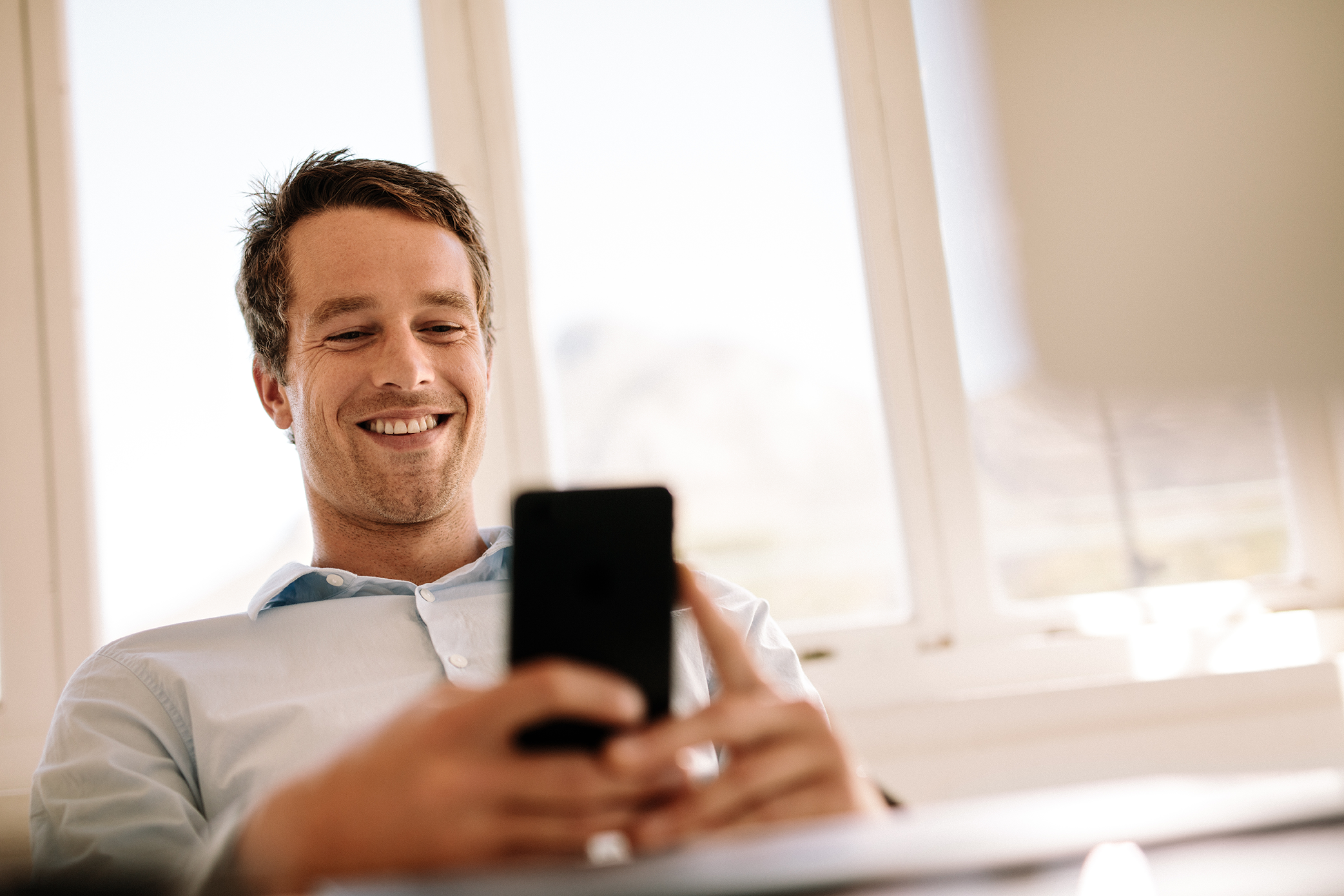 Hombre sonriente sentado en su escritorio utilizando su celular explorando Mobile Device Management un gran aliado para el hombre office.