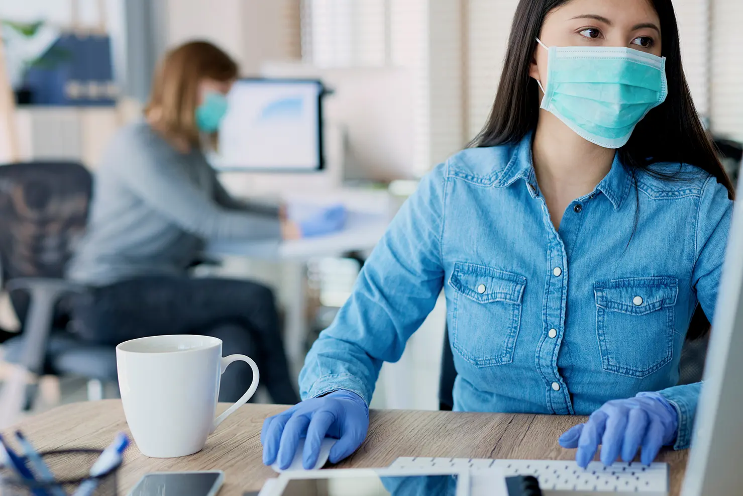 Dos mujeres en oficina frente a escritorio con cubrebocas y guantes de látex, una en primer plano y otra de fondo en segundo plano, mostrando cómo minimiza el riesgo de contagio de COVID-19 en la oficina