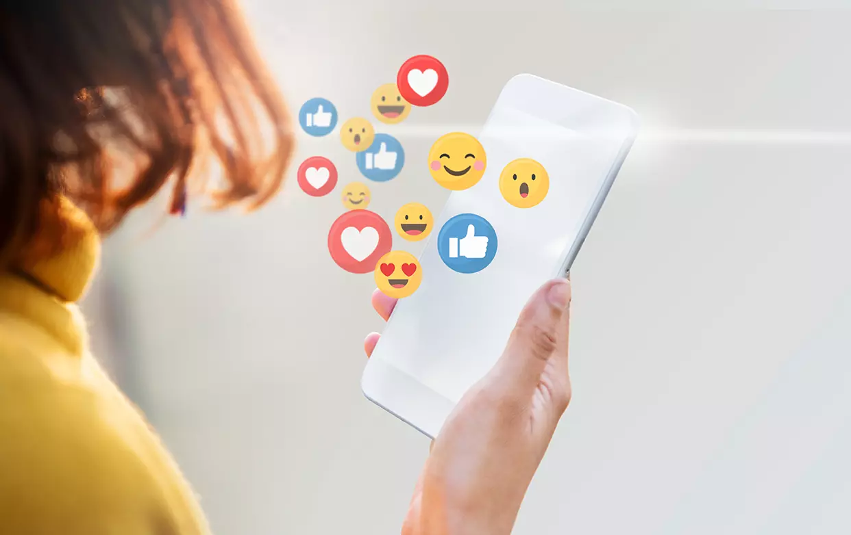 Mujer utilizando su celular con emojis saliendo de el mostrando que Facebook e Instagram anuncian herramientas para combatir adicción a las redes sociales.
