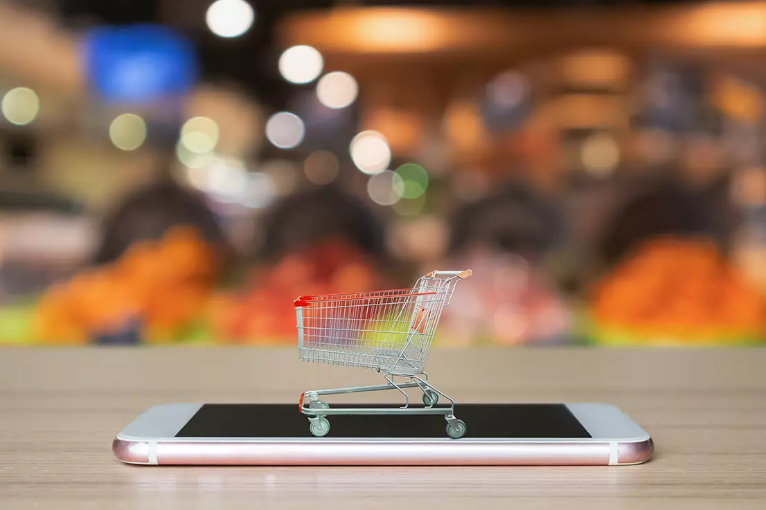 celular sobre una mesa y arriba de él, un carro pequeño de supermercado, representando que Walmart permite ahora pedir el super a través de WhatsApp