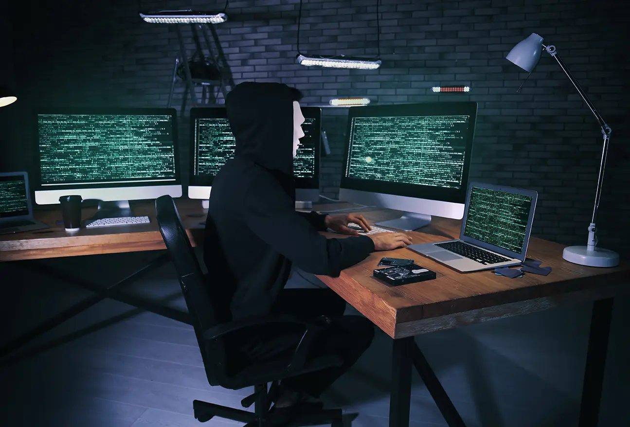 Hacker con capucha frente a una computadora, con varias computadoras de fondo, representando los 7 de los ciberataques más famosos de la historia.